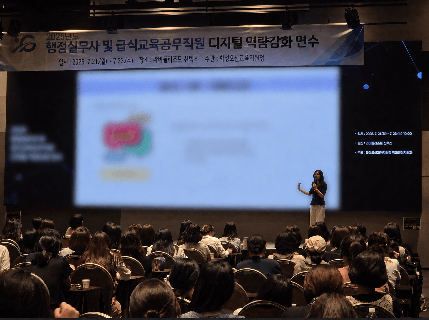 대강당 무대에서 마이크를 들고 대형 스크린 앞에 서서 다수의 청중에게 강의하는 모습